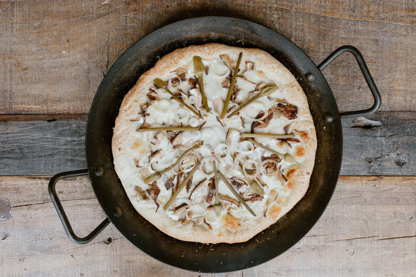 Pickled garlic scapes, chantarelles, onion topped on a pizza in a cast iron pan