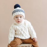 Child wearing the cream Fisherman Sweater and a patterned pom hat, Stone Hollow Farmstead
